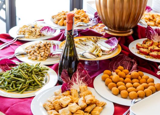 Buffet with vegetables, snacks, and wine on a red tablecloth.