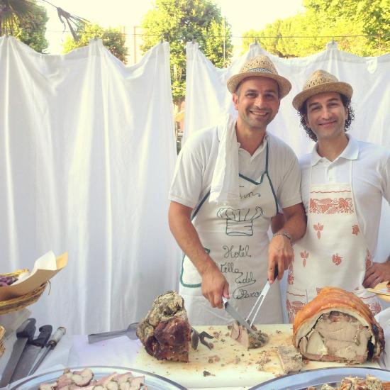 Due uomini servono porchetta con sorrisi e cappelli di paglia.