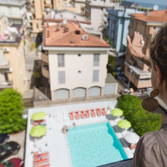 Donna saluta da un balcone, vista su piscina e palazzi.