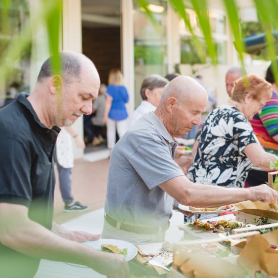 Persone servono cibo a un buffet all'aperto, circondati da piante.