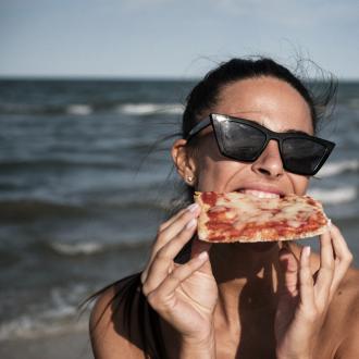 Donna mangia pizza in spiaggia, indossa occhiali da sole.