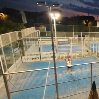 Campo da padel illuminato al tramonto con giocatori e spettatori.