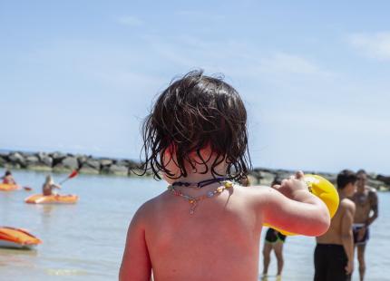 Kind sitzt auf einem roten Tretboot am Strand mit gelbem Ball.