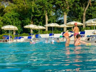 Bambini giocano in piscina con ombrelloni e sedie a sdraio sullo sfondo.