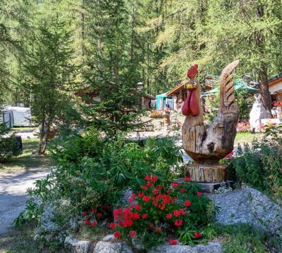 Scultura di gallo in un campeggio tra pini e fiori colorati.