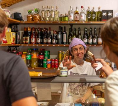 Donna serve clienti in un negozio, scaffali pieni di bevande e snack.