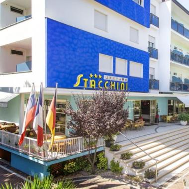 Hotel moderno con terrazza e bandiere, circondato da alberi.