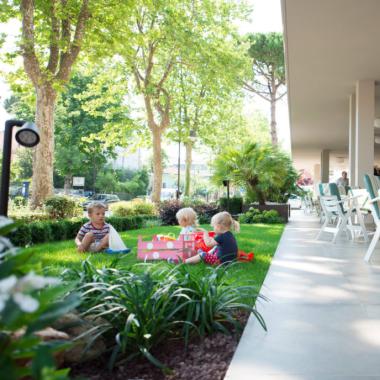 Bambini giocano in giardino accanto a una terrazza con sedie bianche.