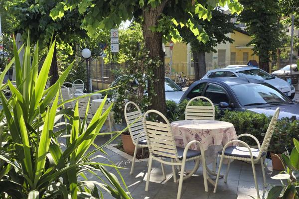 Giardino soleggiato con tavoli, sedie bianche e piante verdi rigogliose.
