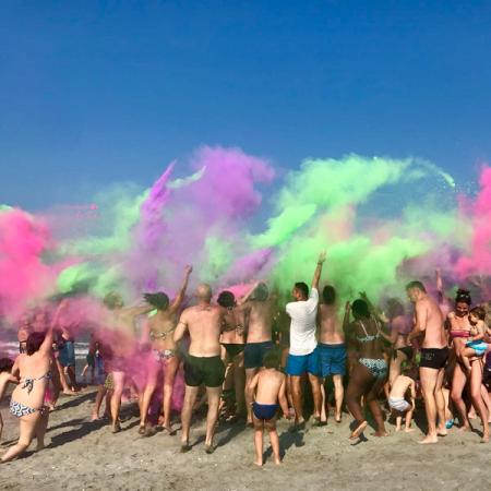 Persone festeggiano in spiaggia lanciando polveri colorate, creando un'esplosione di gioia.