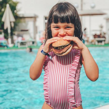 Bambina felice mangia un hamburger vicino alla piscina, indossando un costume a righe.