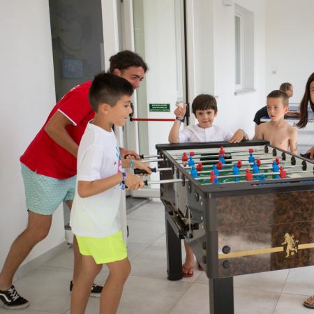 Bambini e adulti giocano a biliardino, sorrisi e divertimento riempiono la stanza.