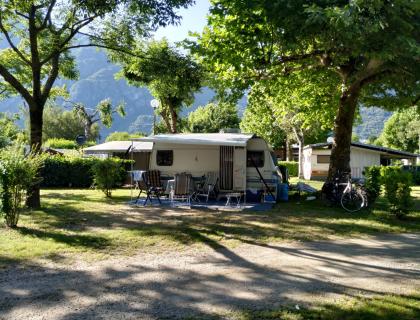 Campeggio soleggiato con roulotte, alberi e montagne sullo sfondo.