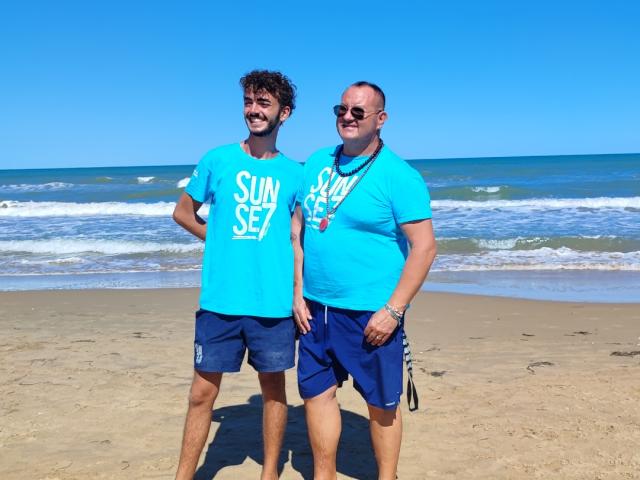 Two men on the beach wearing blue shirts, sea in the background.