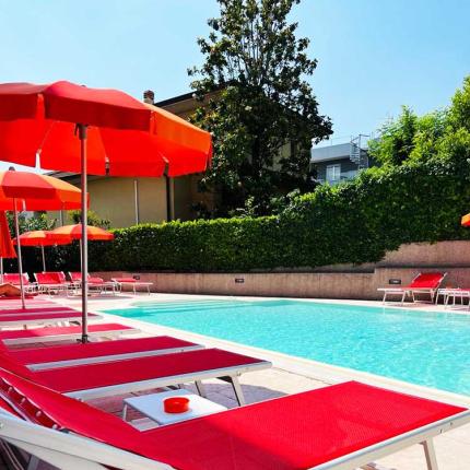 Pool with red loungers and orange umbrellas, surrounded by green hedges.