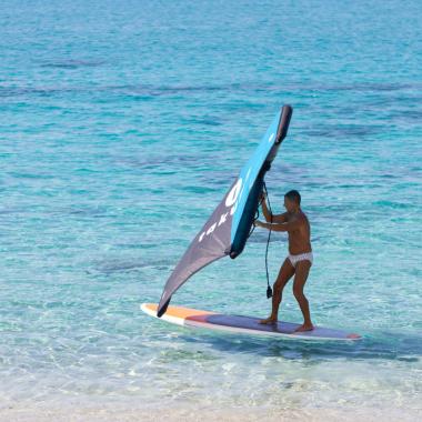 Persona su tavola da surf con vela, mare azzurro.