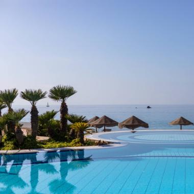 Piscina con vista mare, palme e ombrelloni di paglia.