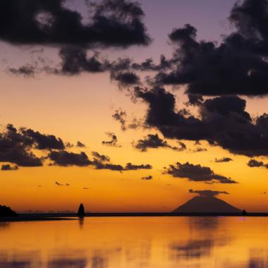 Tramonto su un vulcano con nuvole e riflessi sull'acqua.