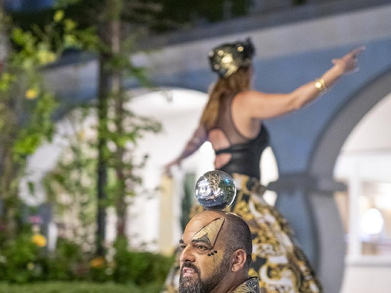 Uomo in abito dorato con sfera sulla testa, donna sullo sfondo.