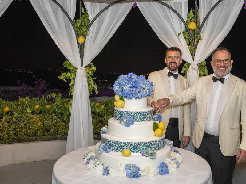 Two men cutting a wedding cake decorated with blue flowers and lemons.