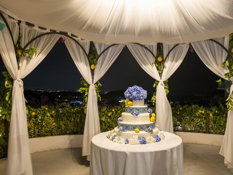 Wedding cake decorated with blue flowers under a gazebo.