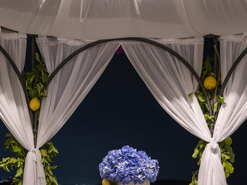 Wedding cake decorated with blue flowers and lemons under a gazebo.
