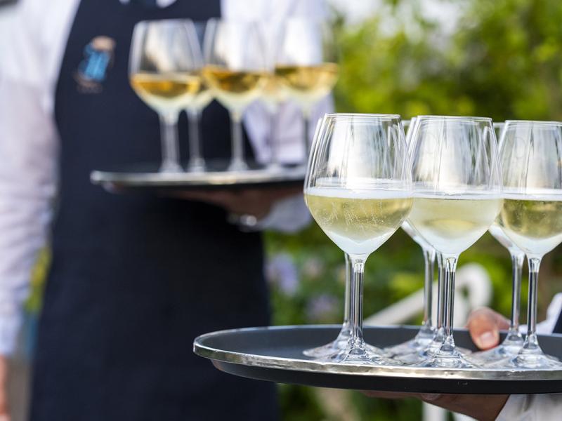 Waiters serve white wine glasses on trays in an outdoor setting.