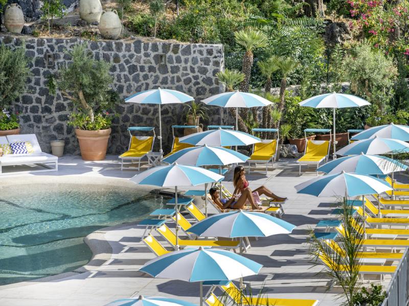 Piscina con lettini gialli e ombrelloni azzurri, circondata da piante.
