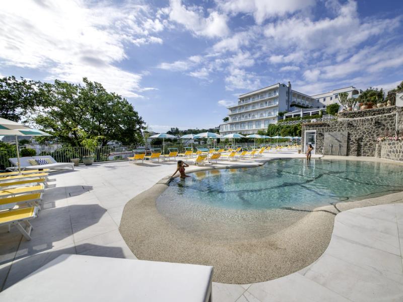 Piscina all'aperto con lettini gialli e ombrelloni, hotel sullo sfondo.