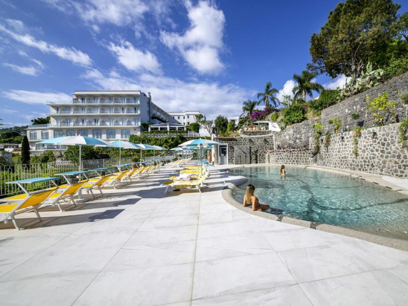 Piscina all'aperto con lettini e ombrelloni, hotel sullo sfondo.
