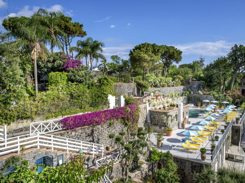 Giardino lussureggiante con piscina e ombrelloni colorati in un ambiente mediterraneo.