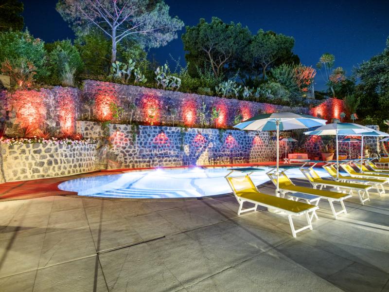 Piscina notturna con lettini e luci colorate, atmosfera rilassante.