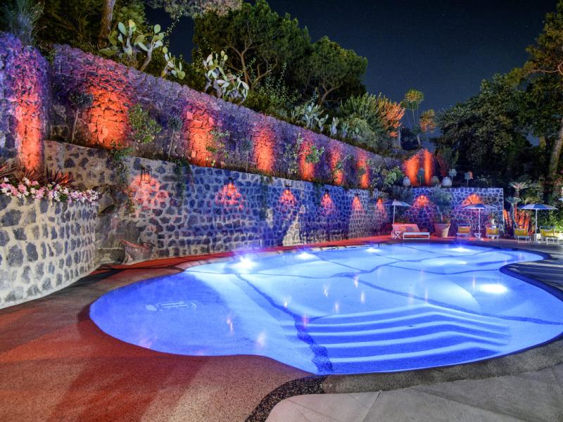 Piscina illuminata di notte, circondata da mura di pietra e vegetazione.