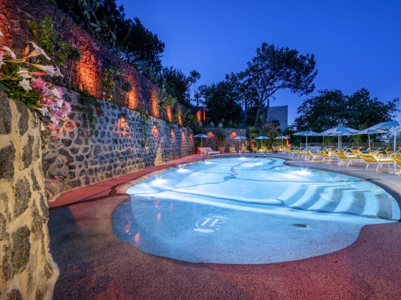 Piscina illuminata di sera con sedie a sdraio e ombrelloni.