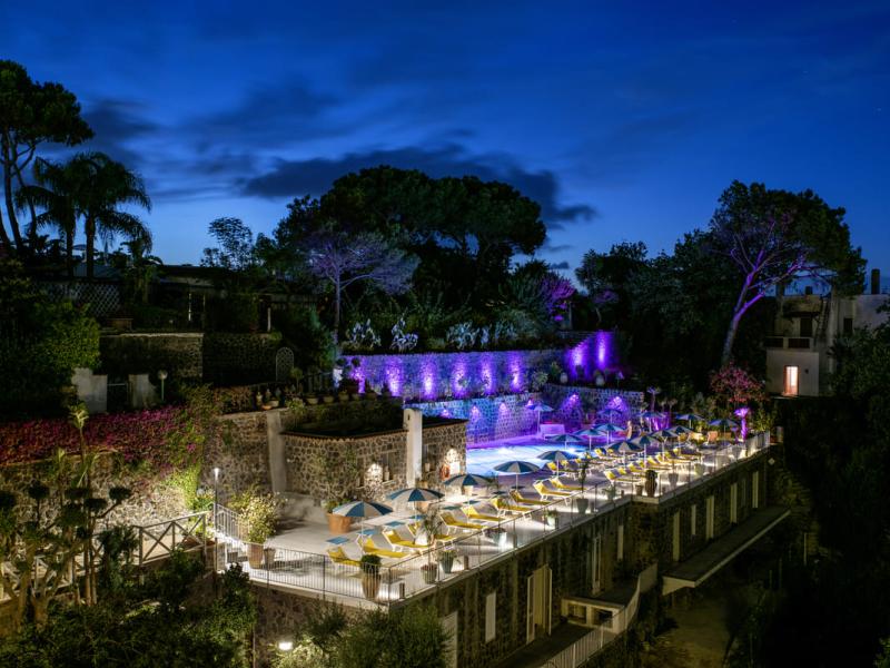 Terrazza illuminata con piscina, ombrelloni e piante in un ambiente notturno.