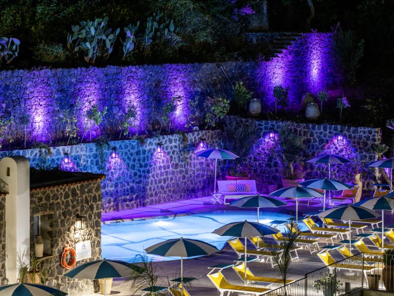 Piscina notturna con luci viola e ombrelloni colorati.