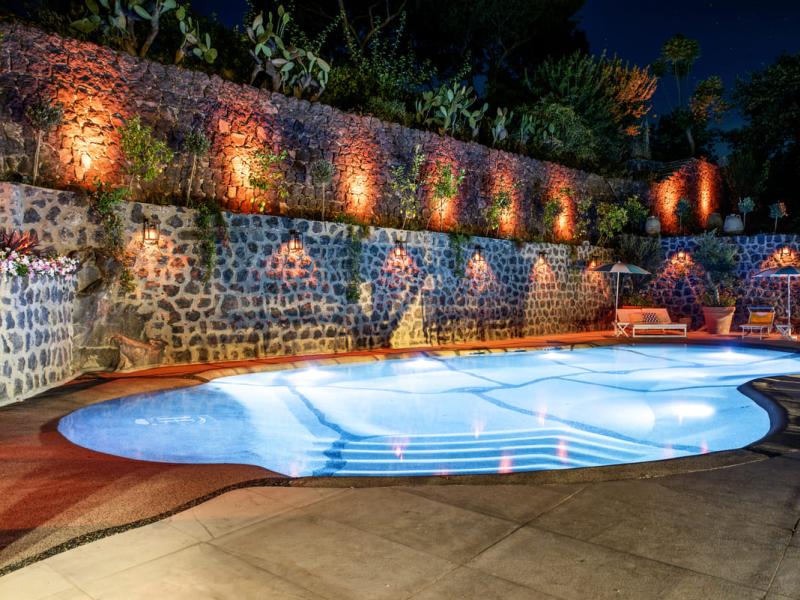 Piscina illuminata di notte con parete di pietra e piante.