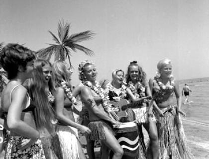 Donne felici danzano in spiaggia, suonando strumenti e indossando gonnellini hawaiani.