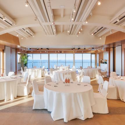 Sala eventi elegante con vista mare, tavoli rotondi e decorazioni minimaliste.