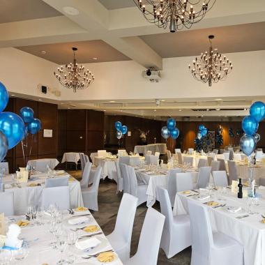 Sala elegante con tavoli bianchi, palloncini blu, atmosfera festosa e calda.
