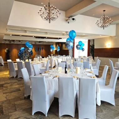 Sala elegante con tavoli bianchi, palloncini blu e lampadari scintillanti.