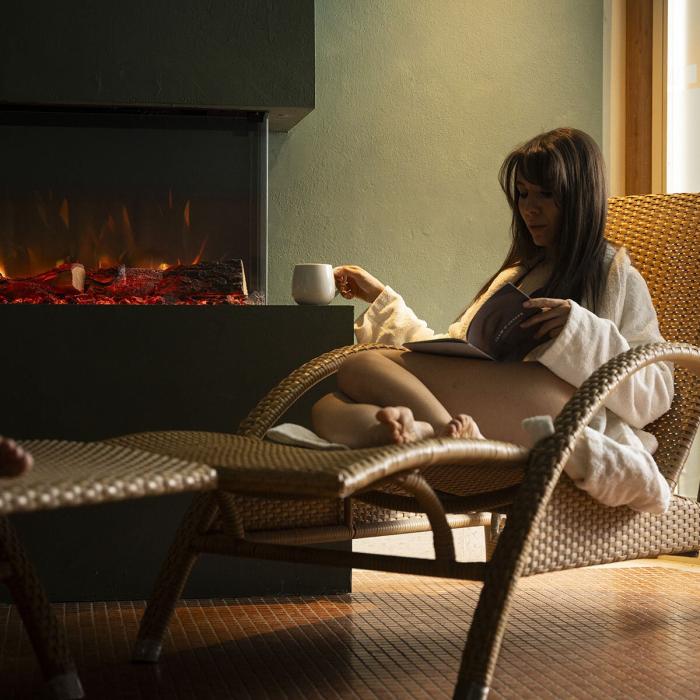 Relaxed woman in chair, sipping tea by a cozy fireplace.