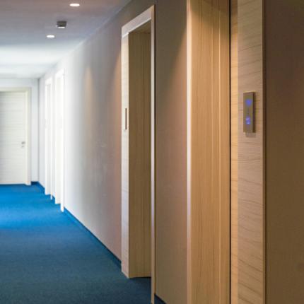 Corridoio d'albergo silenzioso con moquette blu e porte in legno chiaro.