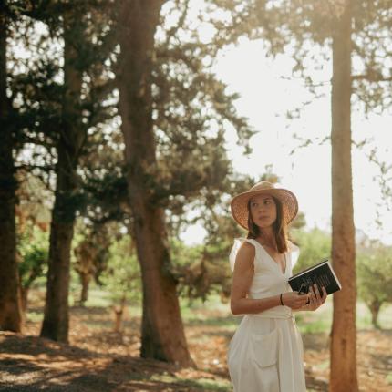 Donna con cappello e libro in un bosco soleggiato.