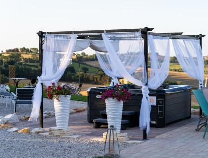 Gazebo con vasca idromassaggio e tende bianche in un paesaggio collinare.