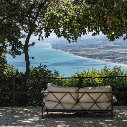Sofa under trees, view of blue sea and countryside, serene and relaxing atmosphere.