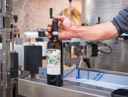 Mano posiziona bottiglia di vino su macchina, evocando artigianalità e cura.