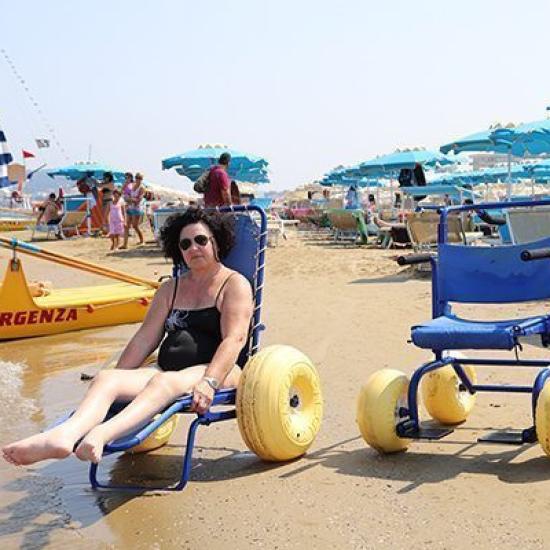 Donna su sedia a rotelle da spiaggia, ombrelloni sullo sfondo.