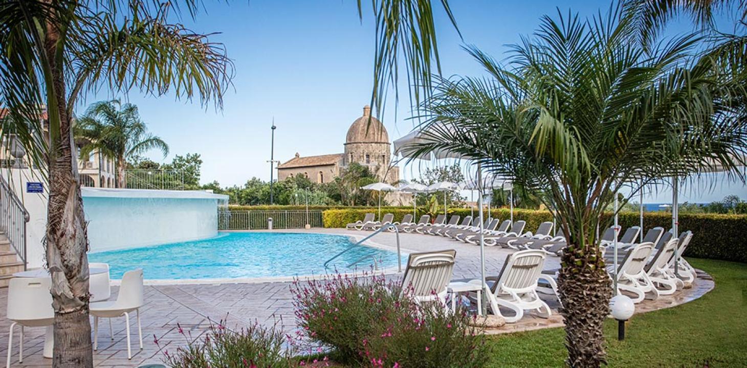 Piscina all'aperto con sedie a sdraio e palme, edificio storico sullo sfondo.