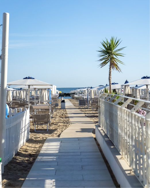 Spiaggia soleggiata con ombrelloni bianchi, passerella e palme.